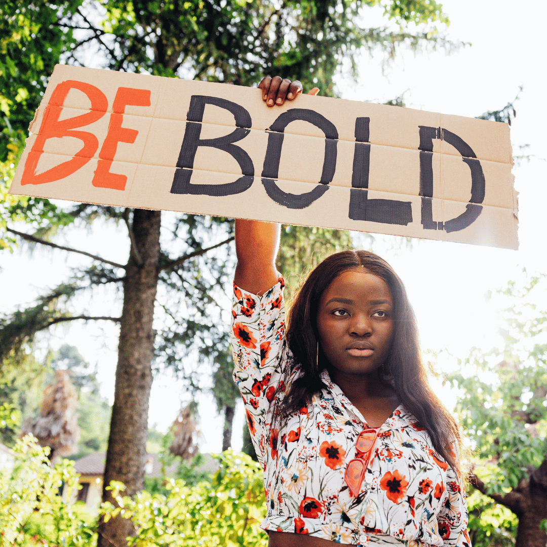 A woman of color holding a sign encouraging women to BE BOLD