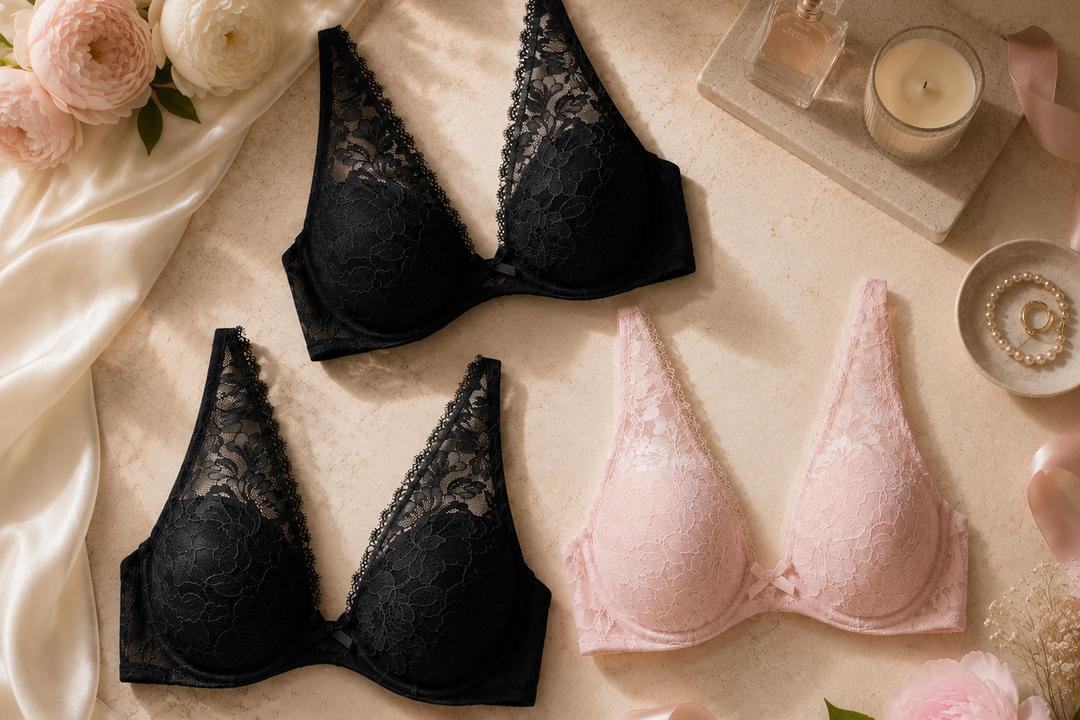 Two lace underwire bras, one black and one light pink, arranged in a styled flat lay with satin fabric, roses, perfume, and a candle.