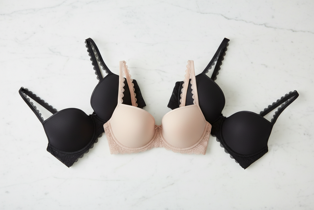 Three underwire t-shirt bras arranged in a row on a white marble surface: two black bras on the sides and one nude beige bra in the center. The bras feature scalloped trim and adjustable shoulder straps.