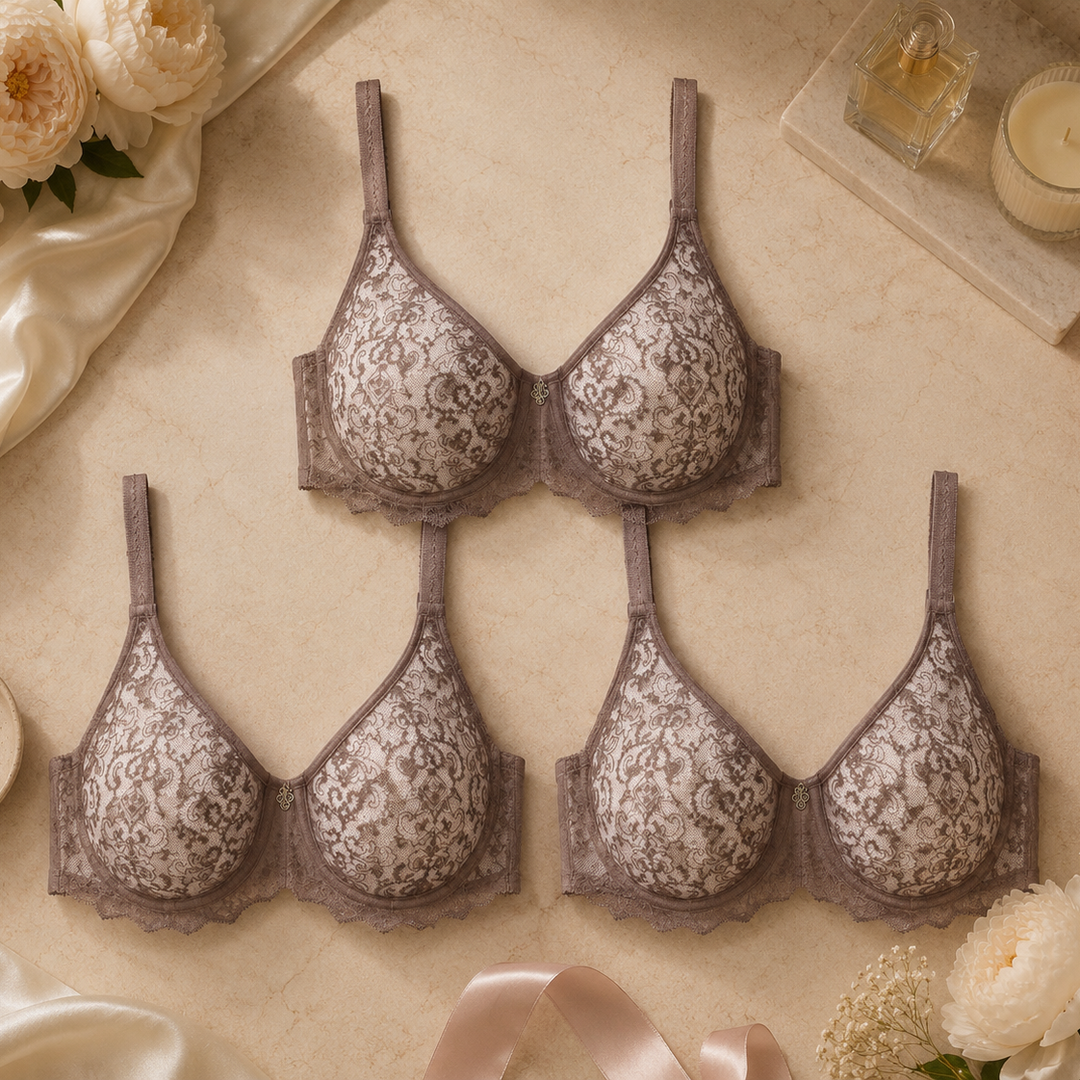 Four lace underwire bras in a muted taupe-gray color are arranged in a flat lay on a beige fabric background, surrounded by cream flowers, satin fabric, a pearl necklace, and a lit candle.