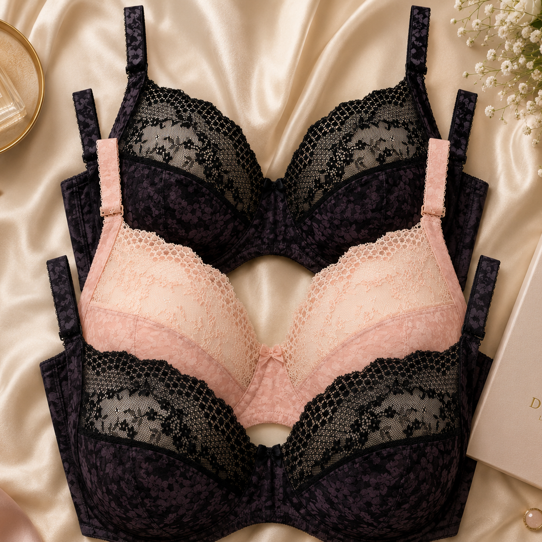 Three large bust plunge lace underwire bras arranged on a satin fabric background, one in black and one in light pink. A perfume bottle, flowers, jewelry, and a branded box are styled around them.