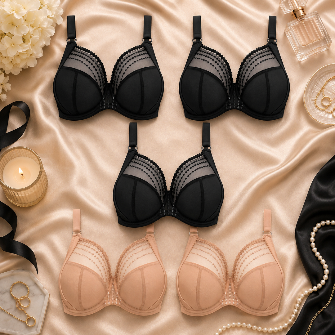 Five large bust underwire bras arranged in a flat lay on champagne satin fabric, with decorative props including a lit candle, pearls, a perfume bottle, flowers, and black ribbon. Two bras are black and two are nude beige.