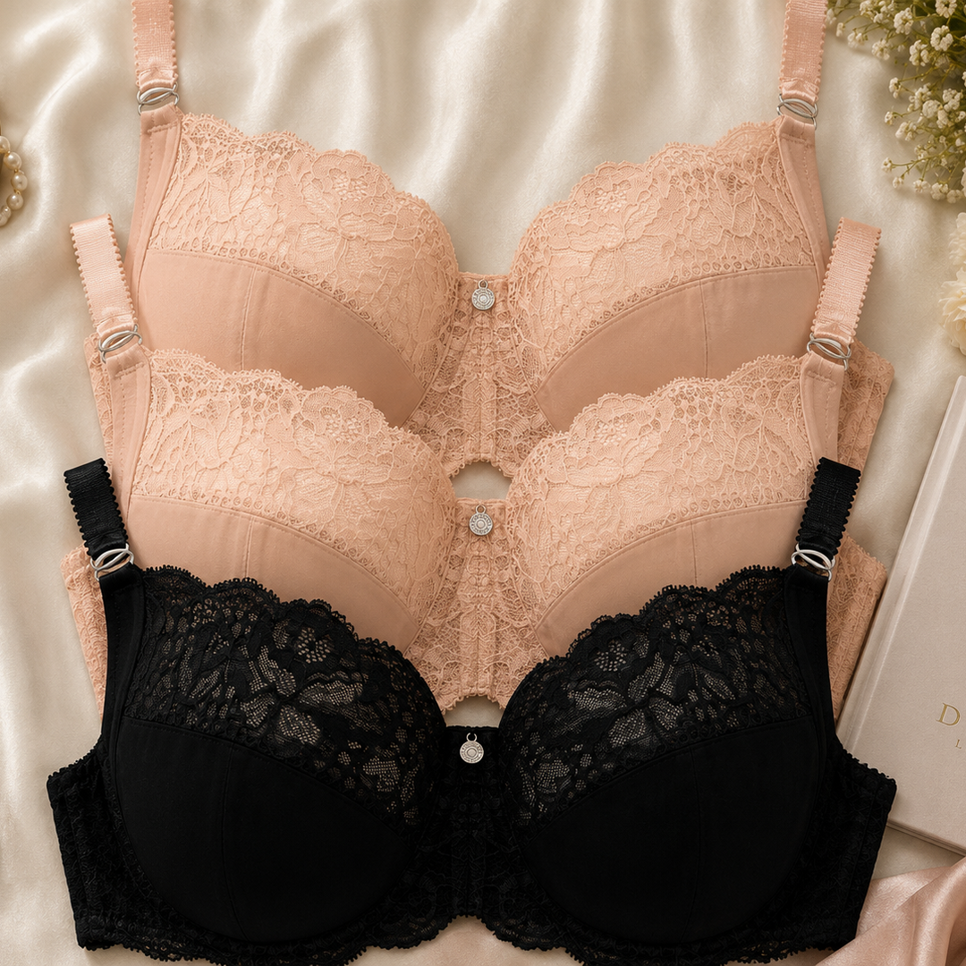 Three lace underwire bras in black, blush pink, and nude beige are arranged in a flat-lay on a white satin fabric with decorative flowers, a perfume bottle, and a book.