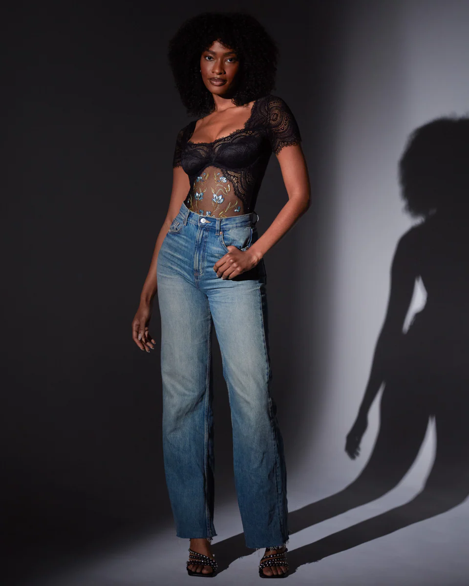 Woman wearing the aster bodysuit and blue jeans standing against a dark background with a shadow on the wall.