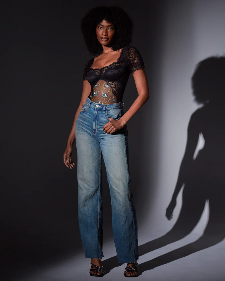 Woman wearing the aster bodysuit and blue jeans standing against a dark background with a shadow on the wall.